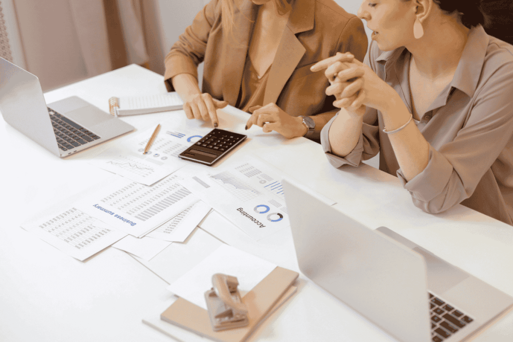 Two accountants sit at a desk in front of their laptops.