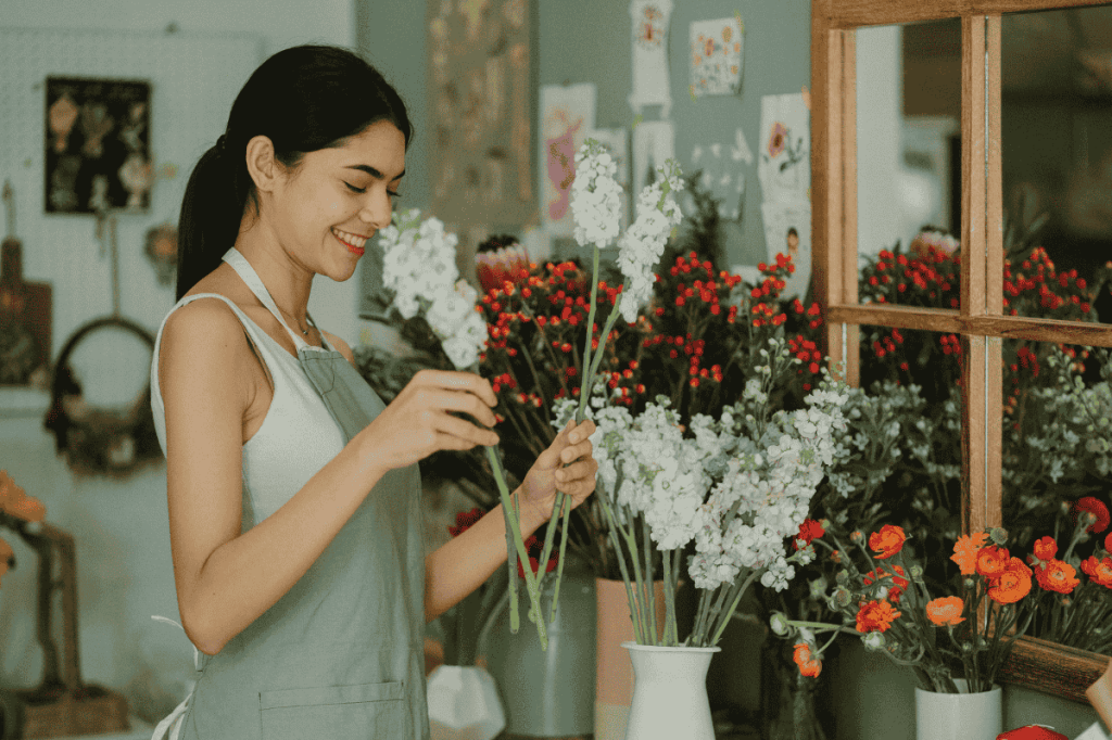 A smiling entrepreneur smiles as she rearranges flowers in her flower shop.