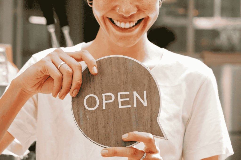 A smiling woman stands holding an open sign in front of her small business.